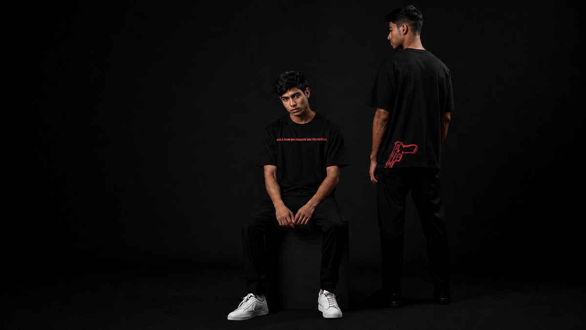 Two models in black t-shirts and black pants posing against a solid black studio background. The seated model wears white sneakers and has red text across the chest, while the standing model shows the back of the black t-shirt featuring a red graphic design.